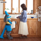A life-sized Lucario holds a dish towel as a woman does the dishes.